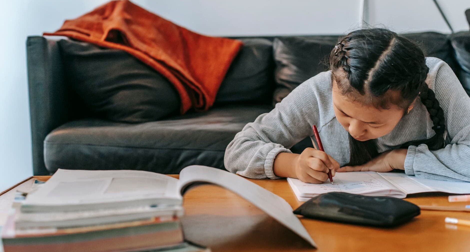 A child bent over a workbook, writing intently at a table, with a stack of textbooks in the foreground