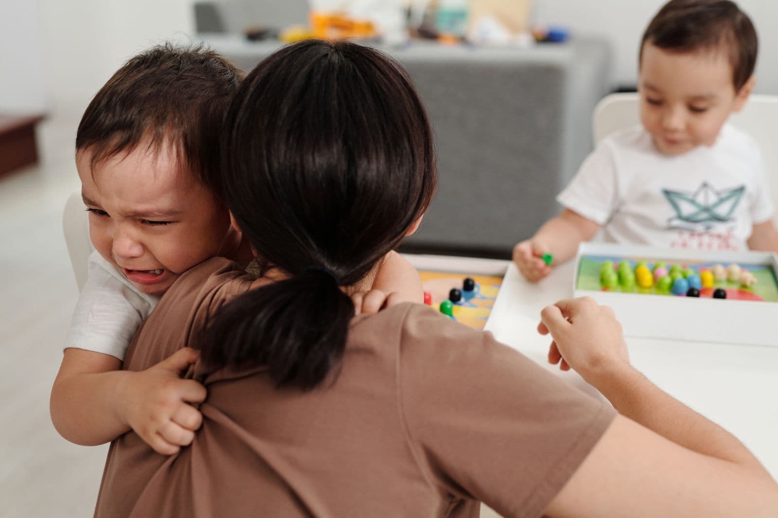 A mother seen from behind holds a crying toddler on her shoulder while a second young child plays with a toy nearby