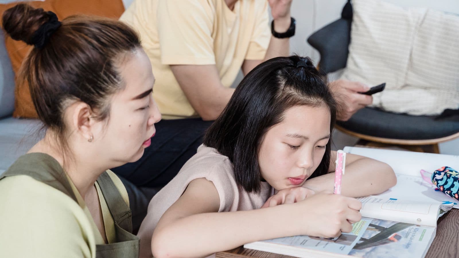 A mother watches closely as her daughter works in an open school workbook at home.