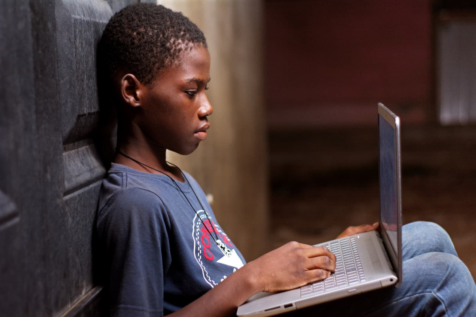 A boy sitting against a wall, focused on a silver laptop screen