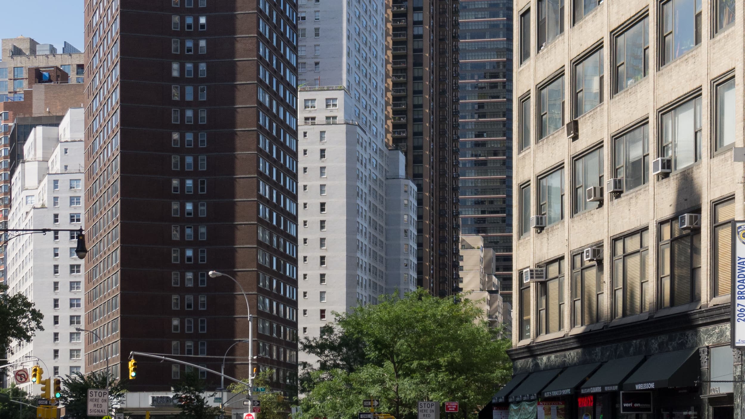 An Upper West Side, Manhattan street scene with residential apartment towers, storefronts, a street tree, and traffic signals under a blue sky.